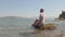 Relaxed woman in white wet tunic sitting on stone in sea waves. Happy tourist woman sitting on rock in sea water and