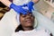 Relaxed Young African Woman Receiving Forehead Massage In Spa. Woman massagist in blue rubber gloves performing facial