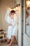 Relaxed young female and male wears white bathrobe, embrace each other, feel relief after taking bath, stand in hotel