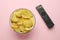 Remote control and bowl of potato chips on pink background, flat lay