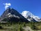 A remote trail leading towards two huge mountains, one covered in snow and one not. There is a glacier in between the mountains.