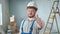 Repairman worker in blue overalls and white helmet is smiling and showing thumbs up. Portrait of redhead man is posing