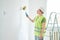 Repairs in the apartment, a man in a green vest and a white helmet stands in a white room with a roller in his hands