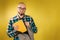 Research and education. A bald bearded man in glasses and a blue plaid shirt puts a book in a backpack. Yellow background. Copy