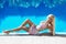 Resort. Beautiful fashion glamour model woman with long wavy hair in luxurious dress with gems relaxing by swimming pool