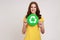 Responsible satisfied teenager girl with wavy hair in yellow t-shirt holding green recycling sign in