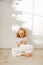 Restful adorable small kid dressed in long white dress, plays with Christmas toy deer, sits crossed legs on wooden floor