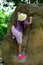 Restless child in panama hat climb up rock in green forest back view. Little girl in comfortable clothes and footwear go