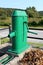 Restored old green outdoor metal water pump with large rotating handle mounted on concrete stand surrounded metal fence and dried