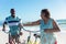 Retired african american senior couple talking while wheeling bicycles at beach on sunny day