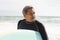 Retired biracial senior man looking away holding surfboard at beach against sky during sunny day