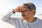 Retired biracial senior man looking away while shielding eyes with hand at beach on sunny day