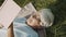 Retired gray haired woman reading a book while resting on the grass in nature