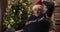 Retired man with gray hair sitting on chair near the Christmas tree. He is wearing warm holiday sweater and Santa hat