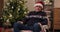 A retired man sits in chair and looks away. He is wearing warm holiday sweater and Santa hat on head. Behind is