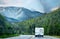 Retro camper driving on highway sucrrounded by evergreen forests and tall mountains with snow patches in distance on overcast day