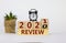 Review 2021 symbol. Fliped wooden cube and changed words `Review 2020` to `Review 2021`. Black alarm clock and house plant.