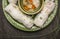 Rice rolls with transparent noodles inside on a green plate with hearbs and vegetable soup on dark wooden rustic background clos