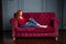 Rich young redhead woman reading a book on red velvet sofa.
