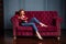 Rich young redhead woman reading a book on red velvet sofa.