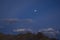 Ridge of gray brown desert mountains under a dark blue evening sky with gray clouds and a full moon with stars