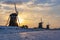 The rising sun is shining just between the blades and the hull of the first windmill on a cold winter morning with a snowy landsca