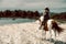 River landscape afternoon. Girl, woman, lady in a work uniform and hat is barefoot. Cowgirl riding pinto horse