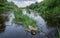 A river with patches of grass and stones of scattered running to the horizon. Long exposure shot. The banks overgrown with lush