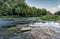 A river with patches of grass and stones of scattered running to the horizon. Long exposure shot. The banks overgrown with lush