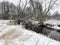 River in Winter: Water in a small river runs through a snow covered prairie with a few bare trees along the banks of the river on