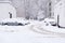 The road is covered with snow. Cars in the adjoining parking lot littered with freshly fallen snow