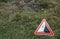 Road sign of falling rocks placed in front of a rocky wall or slope covered partly with vegetation.