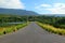 The road stretches along a beautiful river or lake, with mountains, blue sky, white cloud and green forests in the foreground