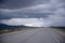 Road stretches into horizon with stormy sky and snow-capped mountains