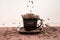 Roasted coffee beans falling down into black cup standing on black plate standing on tablemat.