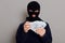 Robber man dressed in black hoodie stands with disguised face and holds a lot of money in his hands, stole a large amount, counts
