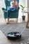 Robotic vacuum cleaner cleaning carpet, woman controls remote control and joy rest while sitting on armchair home