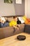 Robotic vacuum cleaner passes to clean the laminate wooden floor, while the young boy playing with tablet computer on the sofa.