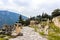 Rock walkway down hill in ancient Delphi Greece past ionic columns and parts of temples and past a reconstructed treasury with