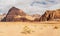 Rocky massifs on red sand desert, few dry grass cluster ground, cloudy sky in background, typical scenery in Wadi Rum, Jordan