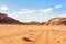 Rocky massifs on red sand desert, vehicle tracks ground, bright cloudy sky in background, typical scenery in Wadi Rum