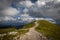 Rocky path surrounded by grass meadow to the top of Klosterwappen, highest peak of Schneeberg, with scenic, cloudy, blue sky