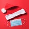 Rolled up Santa Claus hat and disposable medical mask on a red background, top view