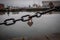 A romantic closed and rusty heart shaped padlock hanging from a chain beside a river with blurred cityscape in the background.