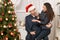 Romantic couple having fun - the girl is sitting on Santa, wants many gifts and makes wishes. Christmas tree with holiday decorati