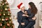 Romantic couple having fun - the girl is sitting on Santa, wants many gifts and makes wishes. Christmas tree with holiday decorati