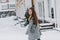 Romantic european girl in long gray coat crosses the street and looking at snowfall. Outdoor portrait of dreamy blonde