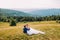 Romantic pair sitting on yellow grass at sunny field with majestic forest hills as background. Back view