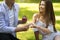 Romantic proposal. Millennial man putting ring on his beautiful girlfriend`s finger in park