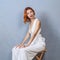 Romantic red-haired woman in white sitting on a wooden chair. Massive jewelry on the ears. Red lipstick. Deep neckline. Hands on